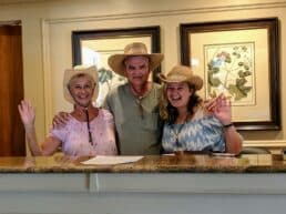 Three smiling individuals wearing straw hats pose together at a reception desk.