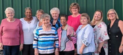 A group of ten women posing together in front of a green background.
