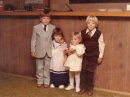Four children in formal attire stand together in front of a wooden counter.
