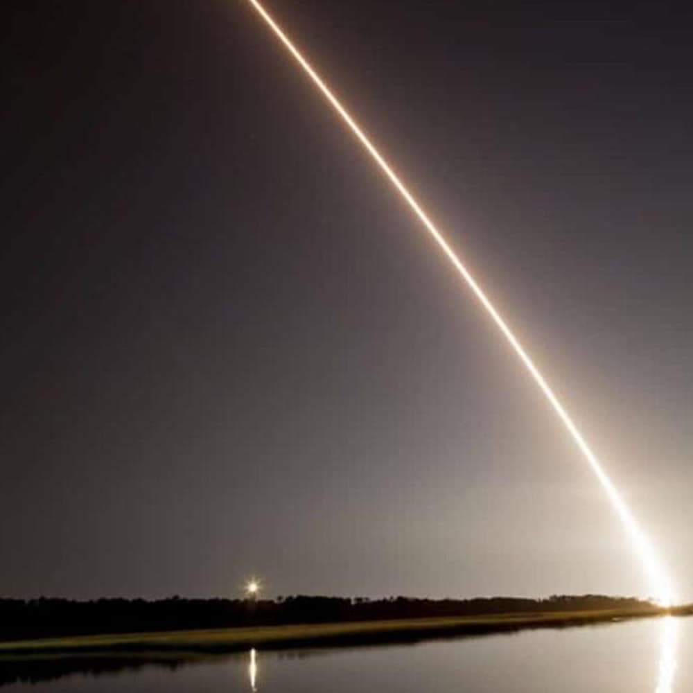 A rocket streaks across the night sky, reflected in calm waters below.
