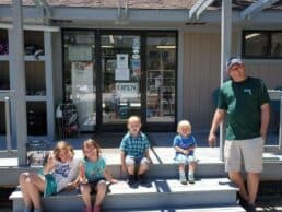 A man and four children sit on steps outside an open shop.
