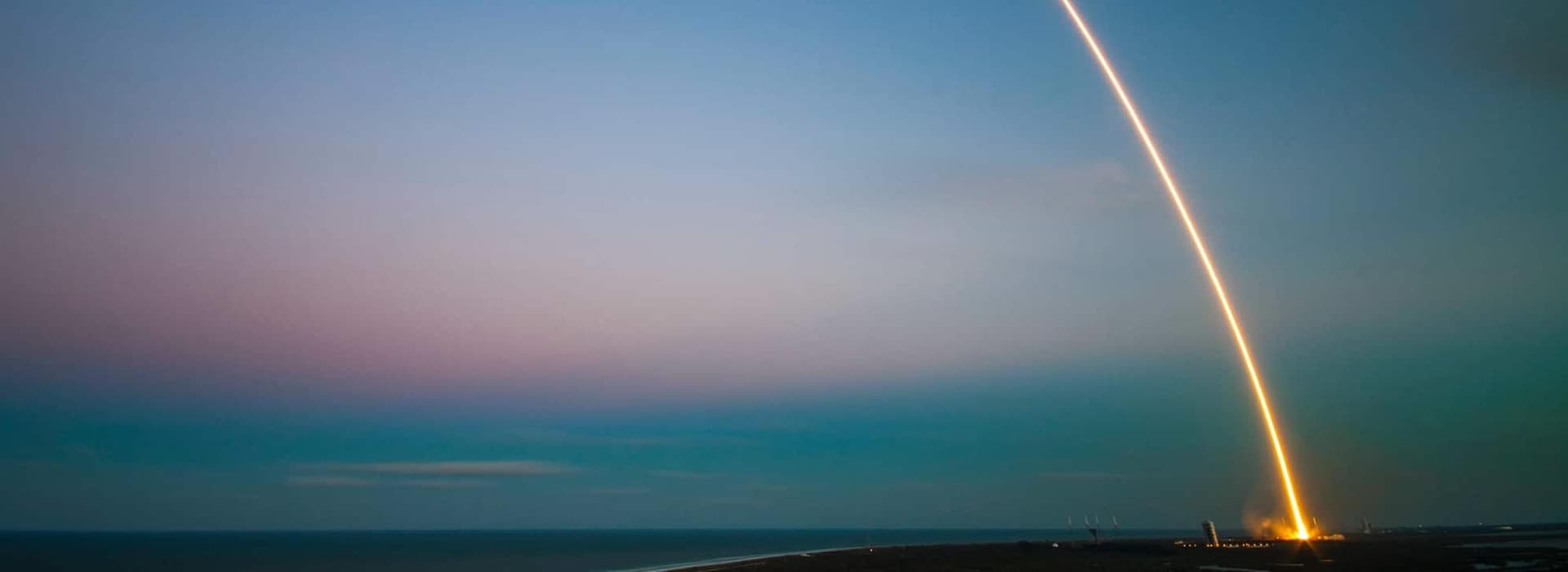 A rocket launches into the sky against a twilight backdrop over the ocean.