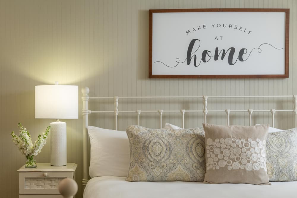 A cozy bedroom scene featuring a decorative sign, plush pillows, a bedside lamp, and fresh flowers.