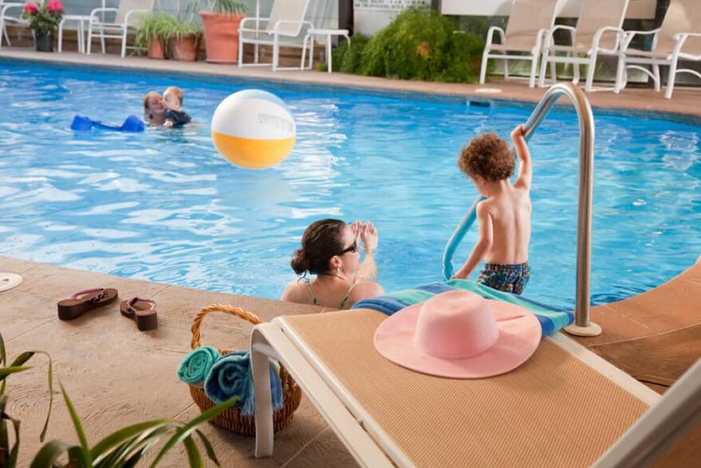 A child climbs the pool ladder while a woman and a toddler play in the water.
