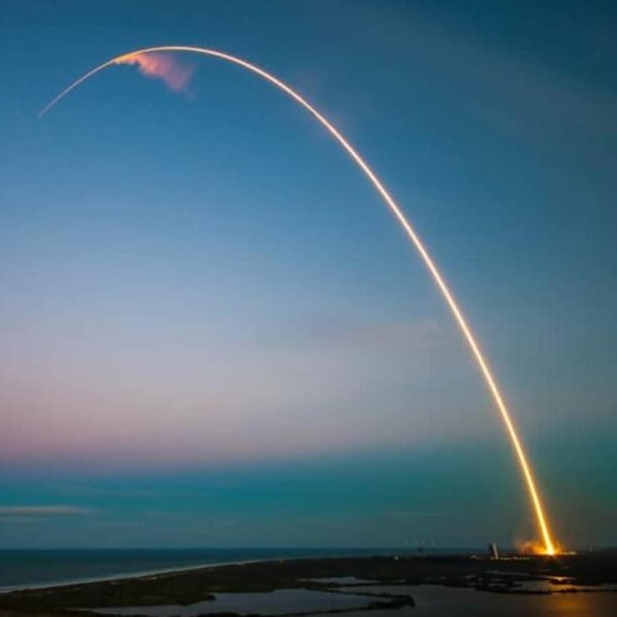 A rocket trail arcs across the evening sky over a coastal landscape.