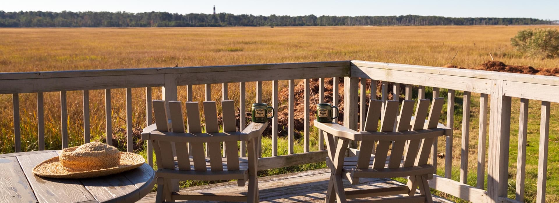 A serene view of a marsh landscape from a wooden deck with two chairs and a straw hat.