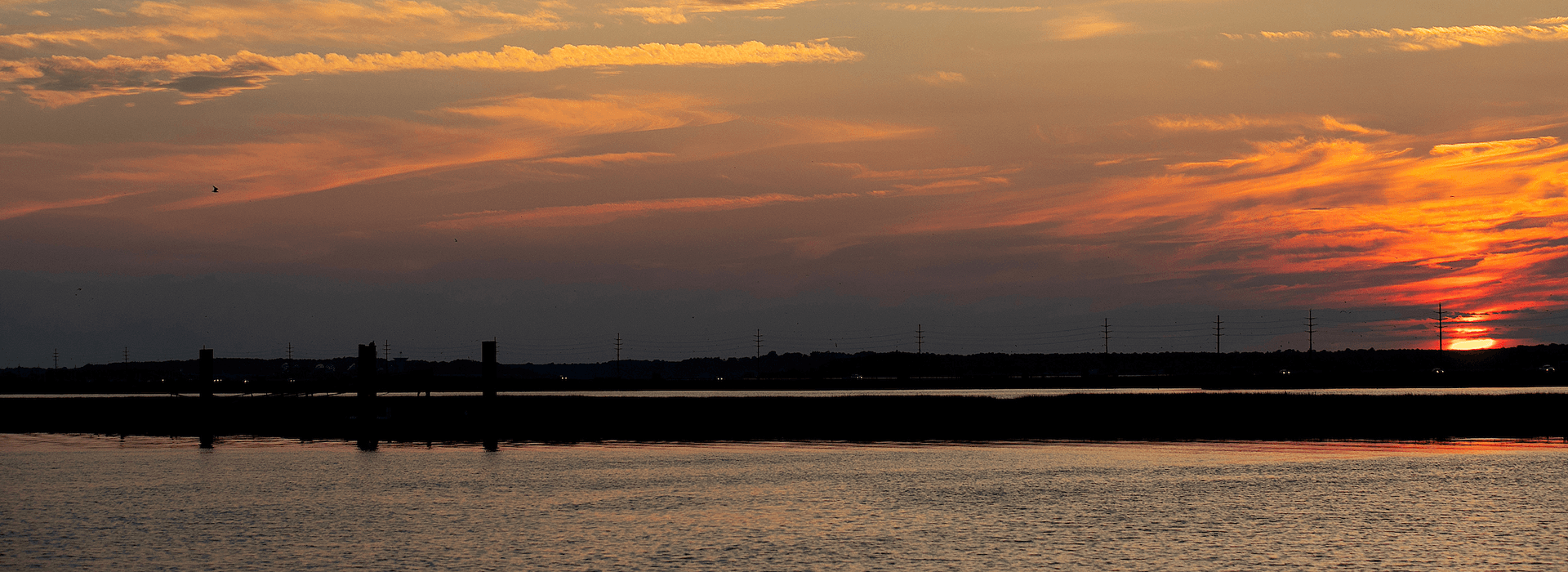 A vibrant sunset reflects on calm water, with silhouettes of distant power lines and poles.