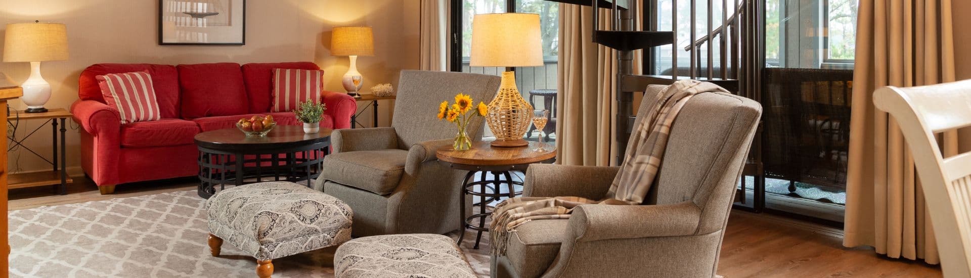 A cozy living room featuring a red sofa, two gray armchairs, a wooden coffee table, and a staircase, with warm lighting and decorative touches.
