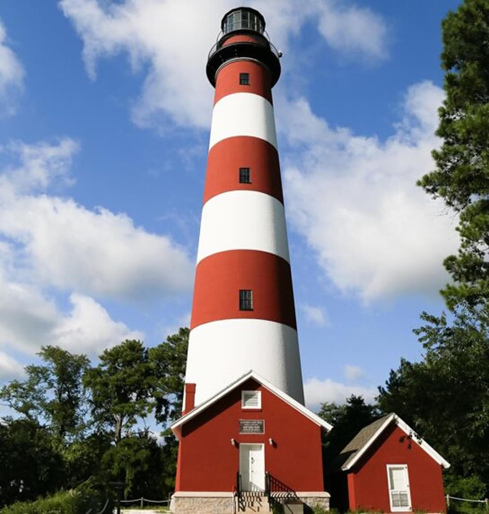A tall red and white striped lighthouse stands next to a small building under a blue sky with clouds.