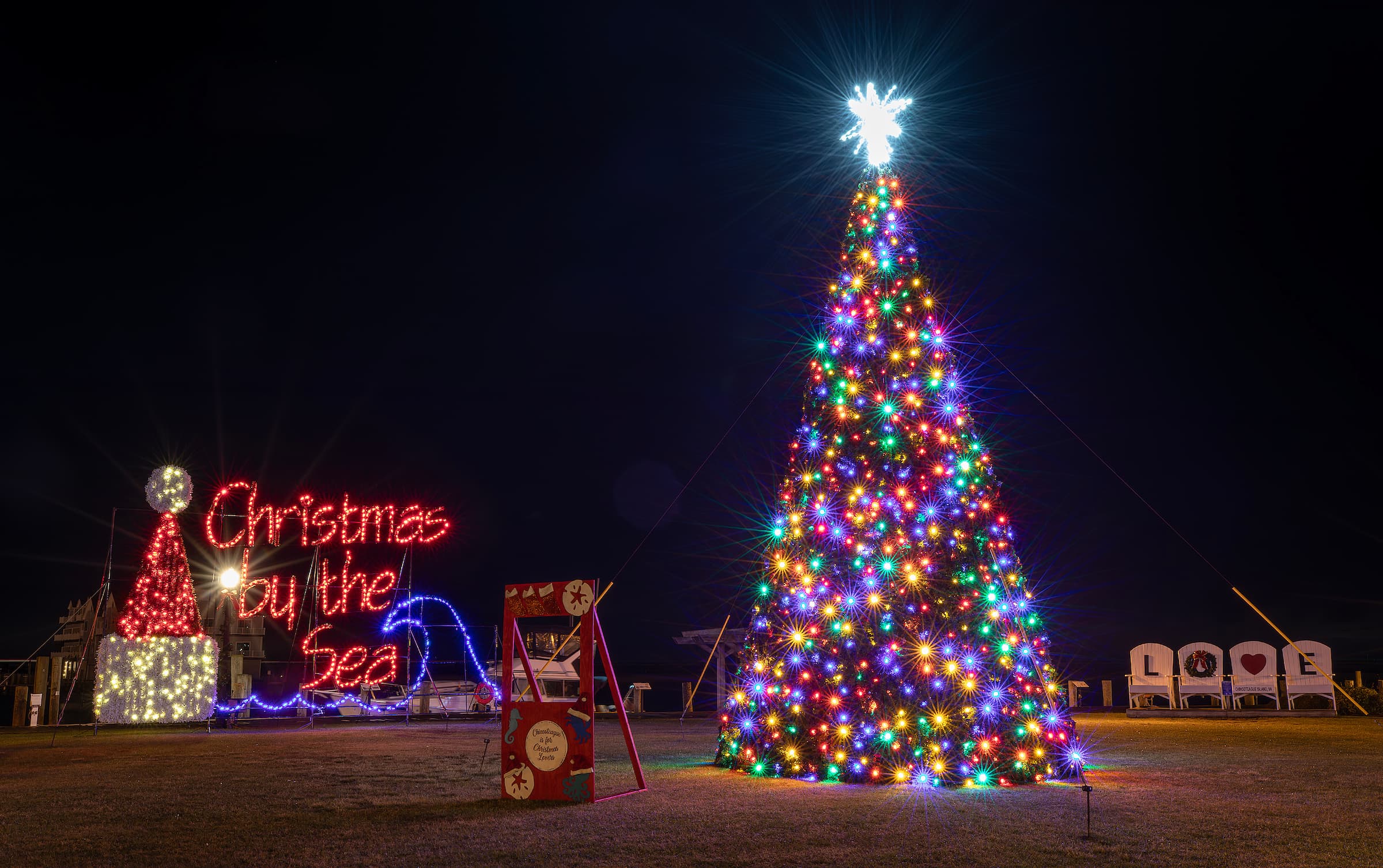A brightly lit Christmas tree stands beside festive decorations that say "Christmas by the Sea" under a night sky. A brightly lit Christmas tree stands beside festive decorations that say "Christmas by the Sea" under a night sky.